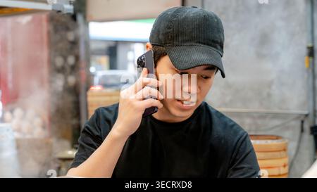 A chef in a bustling Taiwanese restaurant kitchen speaks on the phone while working. He wears a black cap and t-shirt, surrounded by steam and traditi Stock Photo