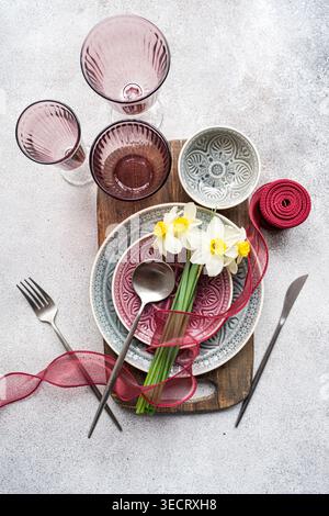 Top view of a spring table setting with stacked plates, cutlery, pink glasses, and a bundle of fresh daffodils tied with ribbon on a rustic wooden boa Stock Photo