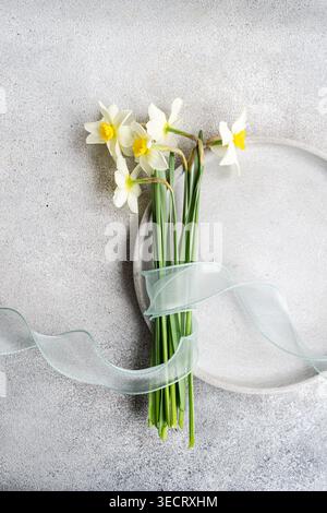 Minimal spring still life with a small bouquet of white and yellow daffodils tied with translucent ribbon beside a ceramic plate on a light gray textu Stock Photo