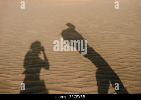 Long shadows of two people fall across rippled sand dunes at sunset in Maspalomas, Gran Canaria, creating a minimalist travel scene with warm light an Stock Photo