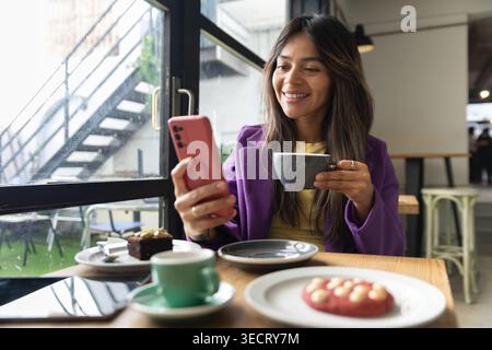 Smiling woman in a modern cafe by a rainy window, holding a coffee cup and checking her smartphone, with desserts on the table during a relaxed coffee Stock Photo