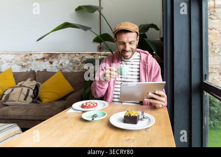 Smiling man in casual outfit enjoys coffee time at a cozy cafe table, sipping a hot drink while browsing on a tablet, with desserts and natural window Stock Photo