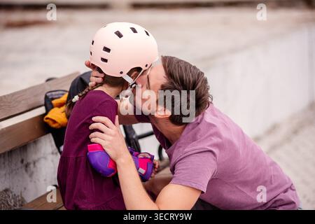 A father kisses his daughter on a bench, both dressed casually. The girl wears a helmet and protective gear, preparing for a roller skating adventure, Stock Photo