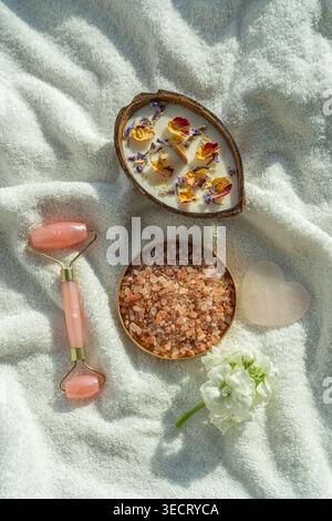 Sunlit spa flat lay on soft towel featuring a coconut shell candle with dried flowers, bowl of spa salt, rose quartz face roller, heart stone, and whi Stock Photo