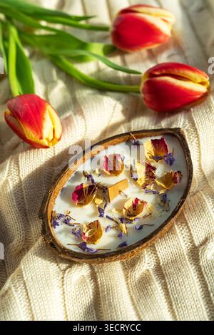 Spa coconut candle in a coconut shell, topped with dried rose petals and blue flowers, resting on a soft knit blanket with red tulips in warm light. Stock Photo