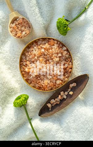 Overhead closeup of pink spa salt crystals in a round bowl with wooden scoop on soft white towel, styled with green flowers for wellness, relaxation, Stock Photo