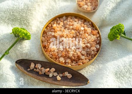 Close-up flat lay of pink spa salt crystals in a wooden bowl on a soft white towel with green flowers, creating a calm wellness, beauty, and relaxatio Stock Photo