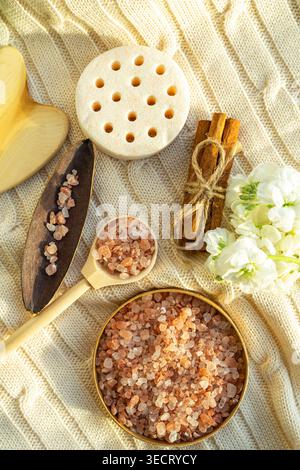 Warm flat lay of spa salt in a bowl with wooden scoop, pumice stone, cinnamon sticks and white flowers on textured beige fabric, creating a calm welln Stock Photo