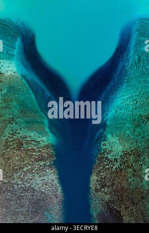 Aerial view of a deep blue lagoon channel cutting through coral reef, turquoise shallows and textured seabed in French Polynesia, creating a striking Stock Photo