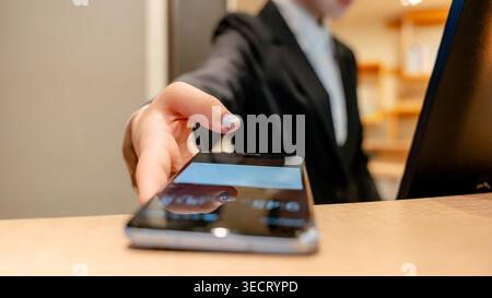 Close-up of a hotel receptionist at the front desk handing a smartphone to a guest for check-in, showcasing Taiwanese hospitality, service, and modern Stock Photo