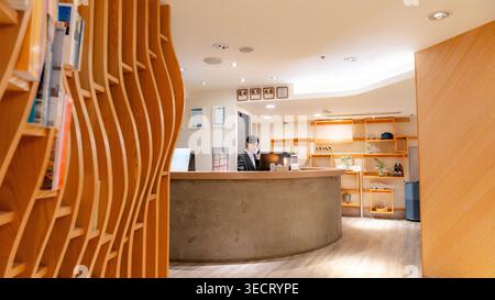 Warmly lit modern hotel lobby with a Taiwanese receptionist working at the front desk, showcasing welcoming hospitality, clean interior design, and tr Stock Photo
