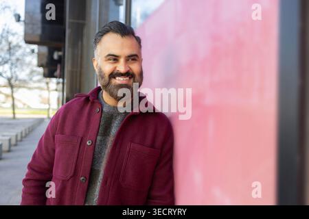 Smiling Arab man with beard wearing a burgundy jacket and sweater, leaning against a modern wall outdoors in the city, looking away in natural dayligh Stock Photo