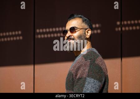 Side profile of an Arab man with beard wearing sunglasses and a warm sweater, standing outdoors beside a modern wall in soft sunlight, confident and r Stock Photo