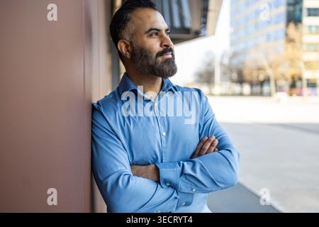 Confident Arab businessman in blue shirt stands with arms crossed, leaning on a wall outside a modern office building, looking ahead with determinatio Stock Photo