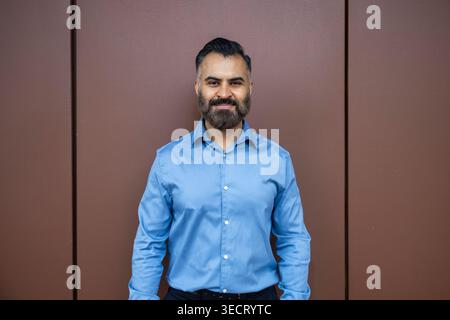 Studio portrait of an Arab businessman in a blue dress shirt, smiling confidently against a simple brown wall background, conveying leadership, profes Stock Photo
