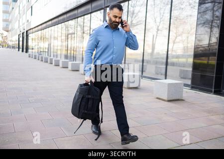 Arab businessman walks briskly outside a modern glass office building while talking on a smartphone, carrying a backpack and dressed in smart business Stock Photo