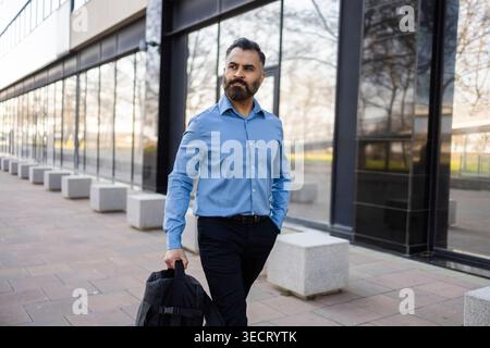 Confident Arab businessman in blue shirt walks beside a modern glass office building, carrying a briefcase, reflecting leadership, professionalism, an Stock Photo