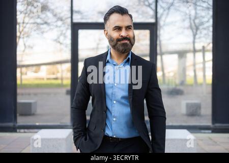 Confident Arab businessman in a dark suit and blue shirt stands outdoors near a modern glass office building, looking away with a thoughtful expressio Stock Photo