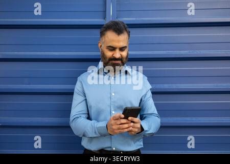 Arab businessman in a light blue shirt stands against a blue metal wall, checking messages on his smartphone. Modern professional lifestyle, communica Stock Photo