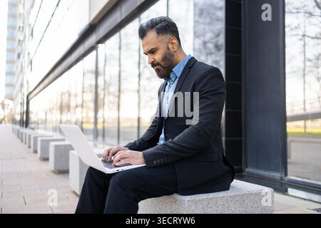 Arab businessman in a dark suit works on a laptop while seated outdoors by a modern glass office building, focusing on remote tasks, communication, an Stock Photo