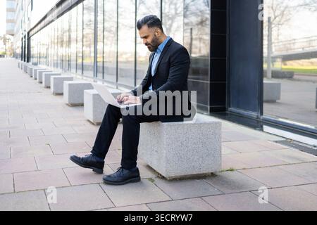 Arab businessman in a black suit sits on a stone bench outside a modern glass office building, using a laptop for remote work, planning, and online co Stock Photo