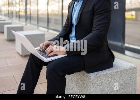 Businessman in a black suit using a laptop while seated on a concrete bench outdoors beside a modern office building, focused on remote work, email, a Stock Photo