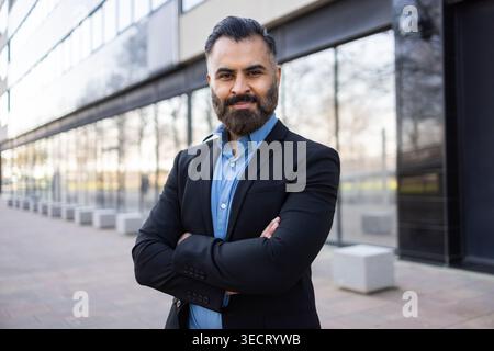 Confident Arab businessman with beard stands with arms crossed outside a modern glass office building, wearing a suit, projecting leadership, success, Stock Photo