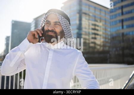 Confident Arab businessman sheikh in traditional keffiyeh and white thobe talking on smartphone outdoors, with modern glass office buildings in the ba Stock Photo