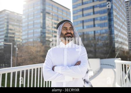Arab businessman sheikh in traditional thobe and keffiyeh stands with arms crossed on an urban bridge, with modern office buildings behind, symbolizin Stock Photo