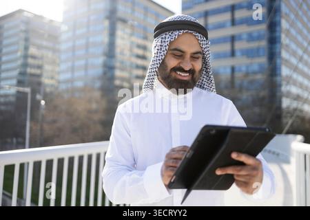 Smiling Arab businessman sheikh in traditional attire uses a digital tablet outdoors near modern office buildings, symbolizing leadership, technology, Stock Photo