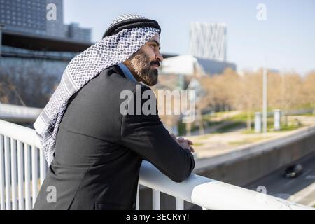 Arab businessman sheikh in black suit and keffiyeh stands on a modern bridge, leaning on the railing and gazing over an urban skyline on a bright day. Stock Photo