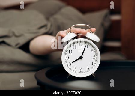 A hand reaches for an elegant white alarm clock resting on a bedside table, capturing the essence of a relaxed day off. The image evokes a sense of ca Stock Photo