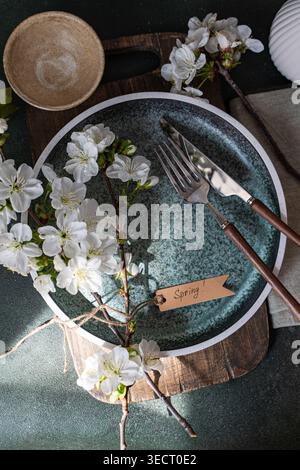 Rustic spring interior table setting with dark ceramic plate, fork and knife, cherry blossom branch, and handmade spring tag on wooden board in soft d Stock Photo