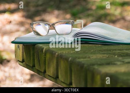 An open book rests on a wooden picnic table with a pair of eyeglasses placed on top. Sunlight filters through the trees, creating a peaceful outdoor r Stock Photo