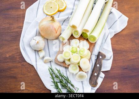 Top view of fresh leeks sliced on a wooden cutting board with kitchen knife, onion, garlic, rosemary and lemon on striped cloth over rustic table, coo Stock Photo