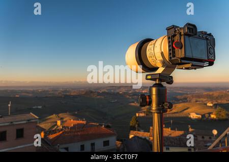 Professional camera with telephoto lens on a tripod overlooking rolling hills and rooftops in Piemonte, Italy, glowing in warm sunset light. Stock Photo