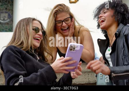Diverse young women laughing together outdoors, sharing a smartphone and enjoying candid social media moments, friendship, connection and joyful, casu Stock Photo
