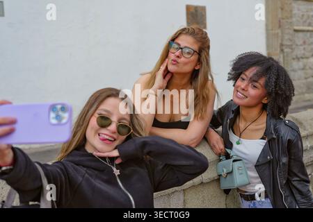 Three young women friends pose for a selfie with a smartphone outdoors, smiling and having fun together, capturing a casual moment of friendship and l Stock Photo