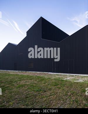 Low angle view of a modern black metal-clad warehouse with sharp geometric rooflines, minimalist doors and walls, set beside a grassy field under a cl Stock Photo