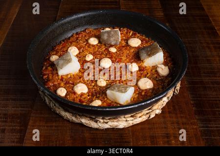 Traditional Spanish rice cooked in a black paella pan, topped with tender cod pieces and creamy aioli, served on a rustic wooden table, ready to share Stock Photo