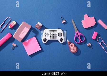Pop art flat lay of school supplies on blue background: pink sticky notes, clips, scissors and tape with a gamepad-shaped calculator, blending Gen Z g Stock Photo