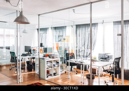 Bright modern office interior with glass partitions, white desks, computers, and ergonomic chairs. Clean workspace with shelves, lamps, and sunlit win Stock Photo