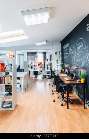 Bright modern open-plan office with long desks, laptops, plants, and a chalkboard wall covered in sketches, creating a creative startup workspace with Stock Photo