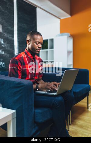 Young Black Man in a red plaid shirt works on a laptop while sitting on a blue sofa in a modern office lounge, focused on remote work, study, or onlin Stock Photo