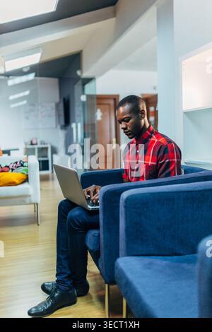 A focused young Black Man in a red plaid shirt sits on a blue sofa, typing on a laptop in a bright modern office lounge with glass walls and cozy seat Stock Photo