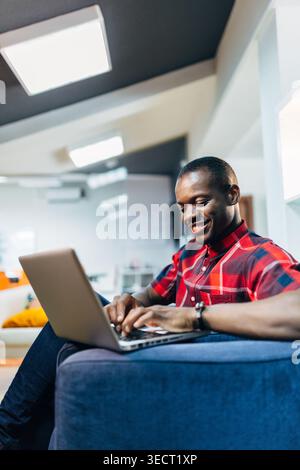 Smiling young Black Man in a red plaid shirt using a laptop on a cozy sofa at home. Relaxed remote work, online learning, and casual browsing in a bri Stock Photo