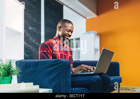 Smiling young Black Man in a red plaid shirt works on a laptop while sitting on a blue sofa in a bright modern living room, enjoying remote work and o Stock Photo