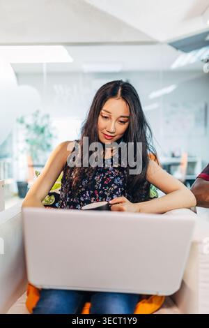 Young woman relaxes on a couch using a laptop and holding a credit card, browsing an online store and making a secure payment. Cozy home interior, cas Stock Photo