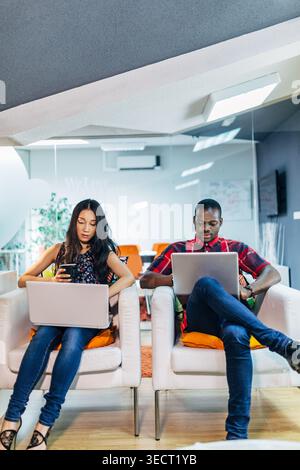 Two young professionals sit in armchairs in a bright modern office lounge, working on laptops and a smartphone, suggesting remote work, productivity, Stock Photo