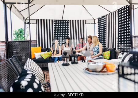 Diverse group of friends sit on a cozy outdoor patio under a gazebo, using laptops and discussing ideas together, with cushions, striped curtains, and Stock Photo